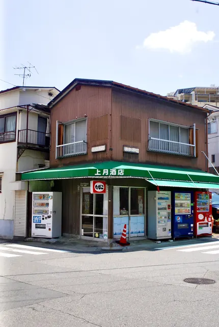 緑の日よけと自動販売機が並ぶ、角の古い酒店