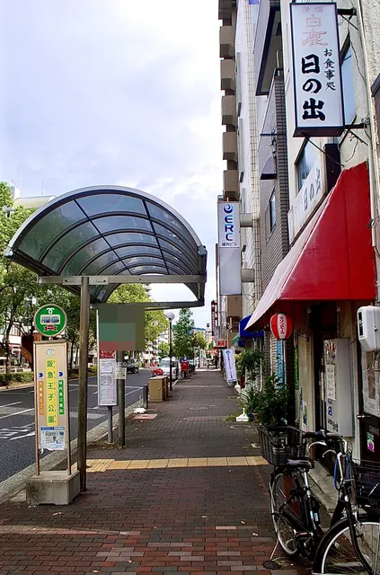「日の出」の看板と赤い日よけ、屋根つきのバス停の歩道