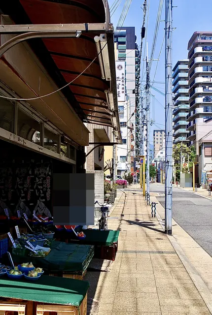 手書きの値札と野菜が並ぶ八百屋の店先と、並木の続く歩道