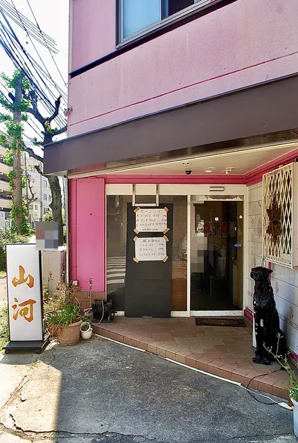 ピンクの壁と「山河」の看板、黒い犬の置物がある喫茶店の入口