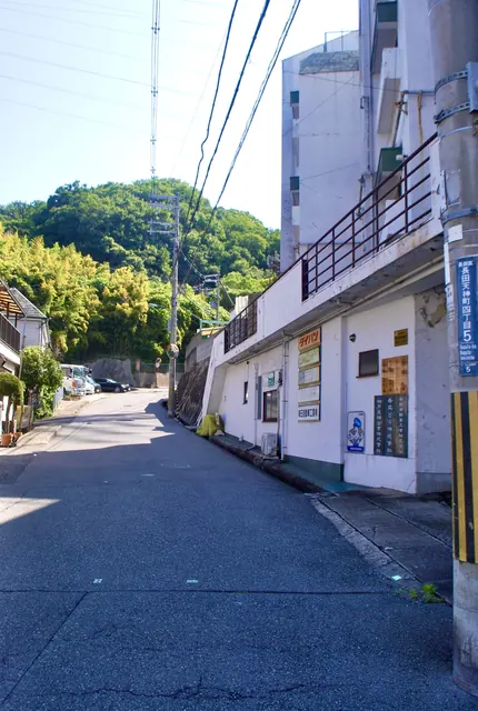 古い看板が並ぶ白い建物と、山へ向かう坂道
