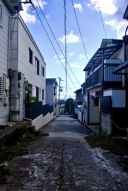 ひび割れた舗装の先に、家々のあいだを奥へ続く細い路地