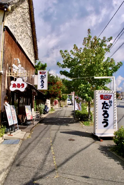 古い木の外壁の店と、大きな木の横に立つ縦長の看板がある道