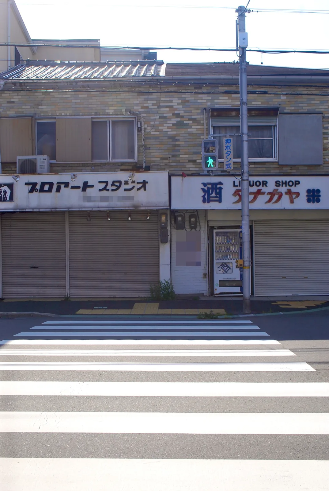 横断歩道の向こうに、シャッターの下りた写真店と酒店が並ぶ風景