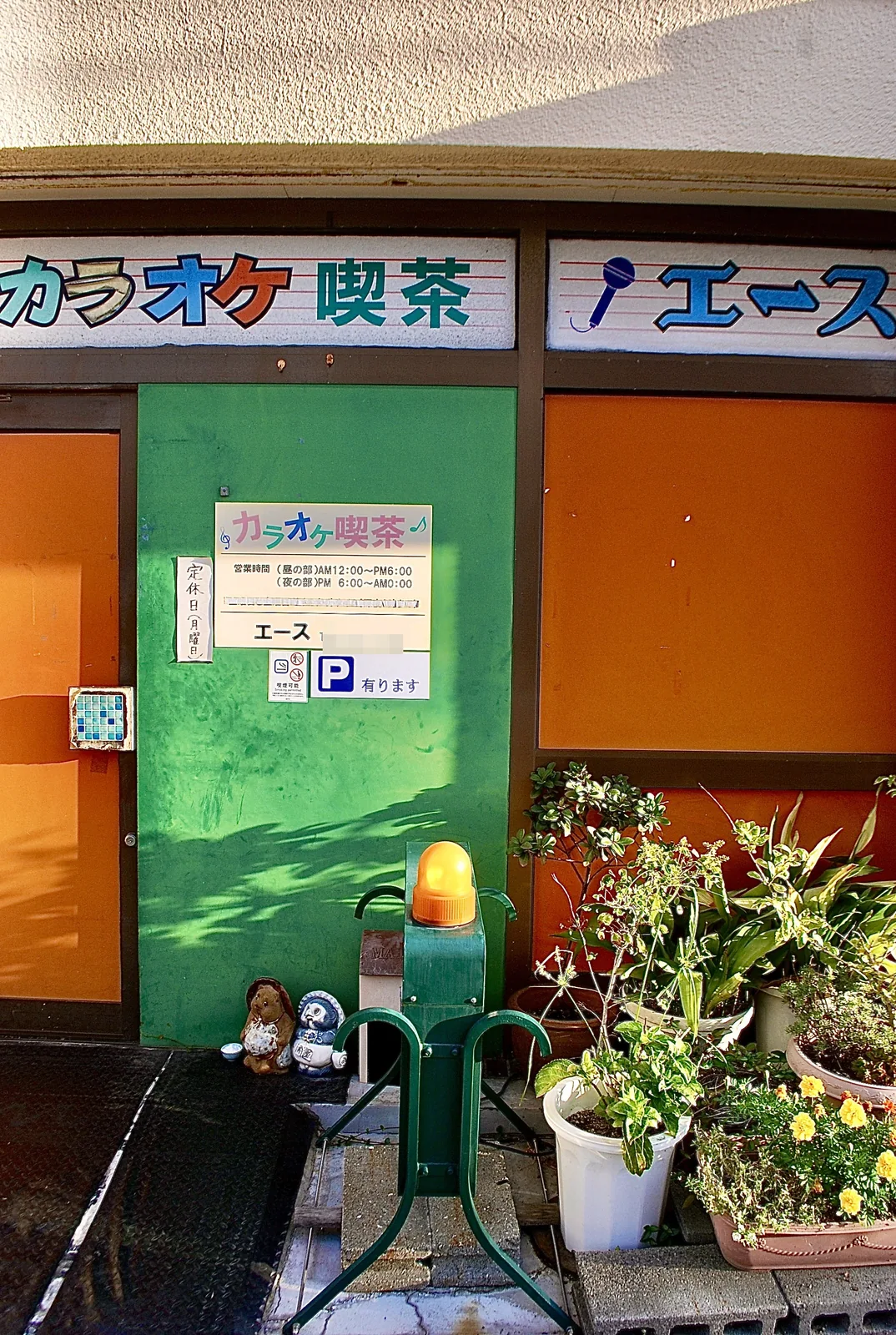 緑の扉とオレンジの窓、植木鉢が並ぶカラオケ喫茶の店先