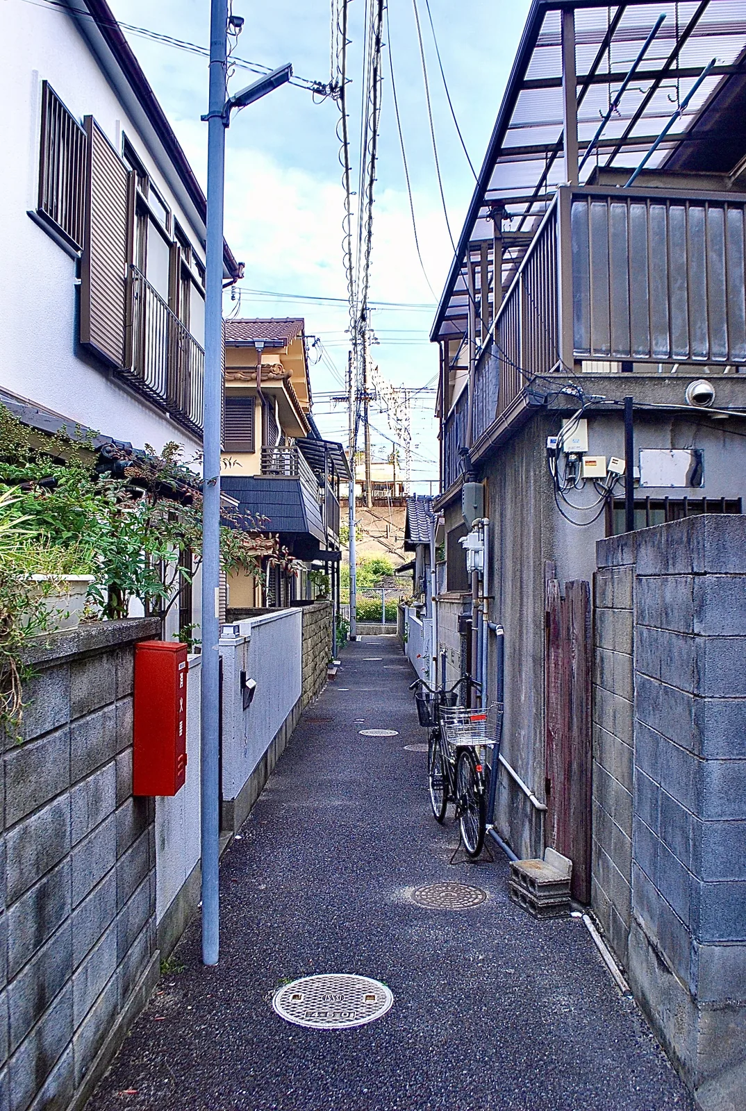 赤い消火器の箱と自転車のある、家々のあいだの細い路地