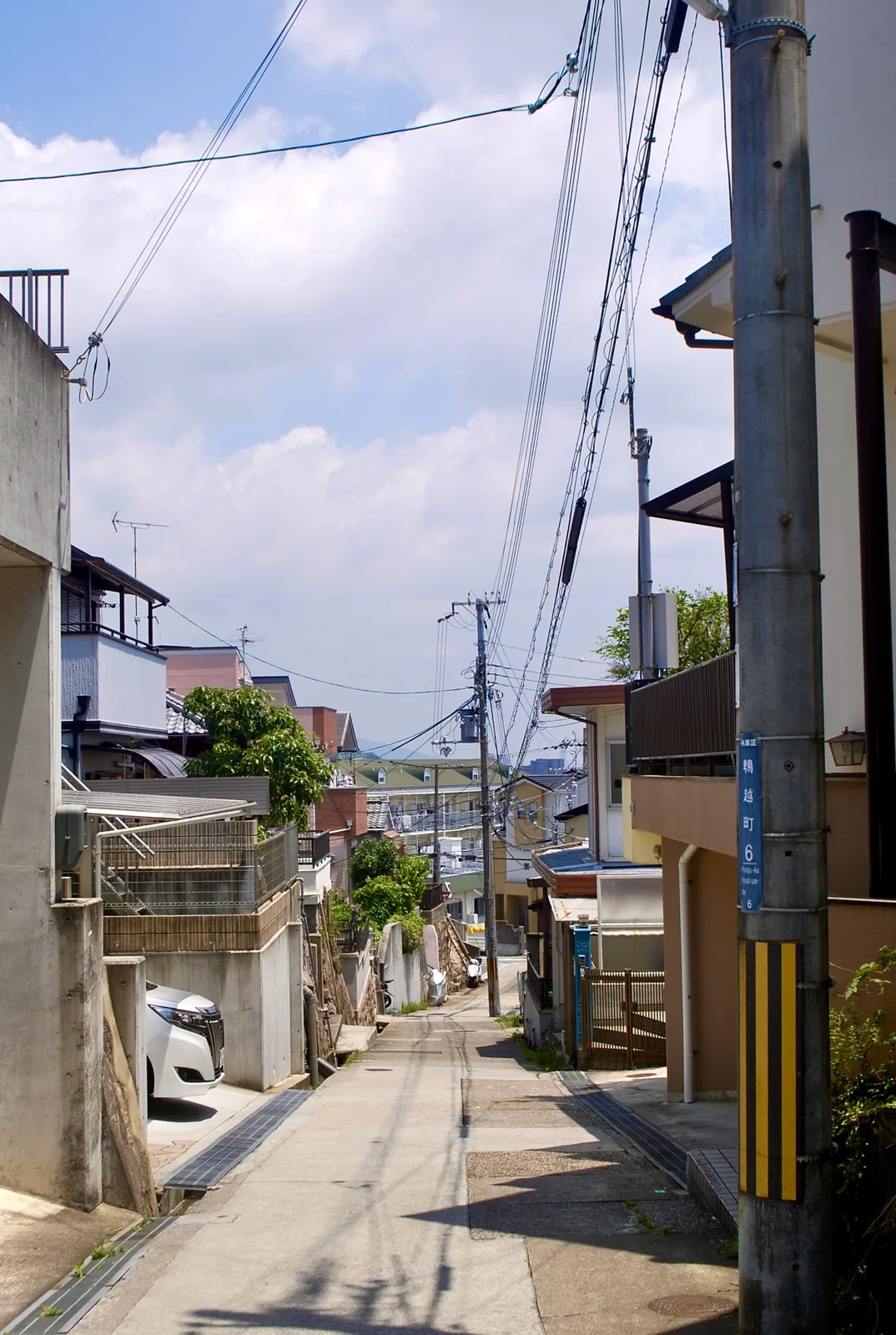 町名表示のある電柱の脇から、遠くの町並みへ下っていく坂道