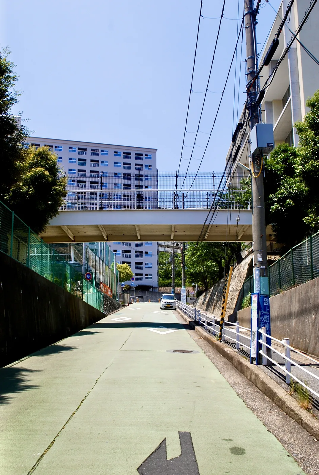緑色に塗られた坂道の先に、歩道橋と団地が見える風景