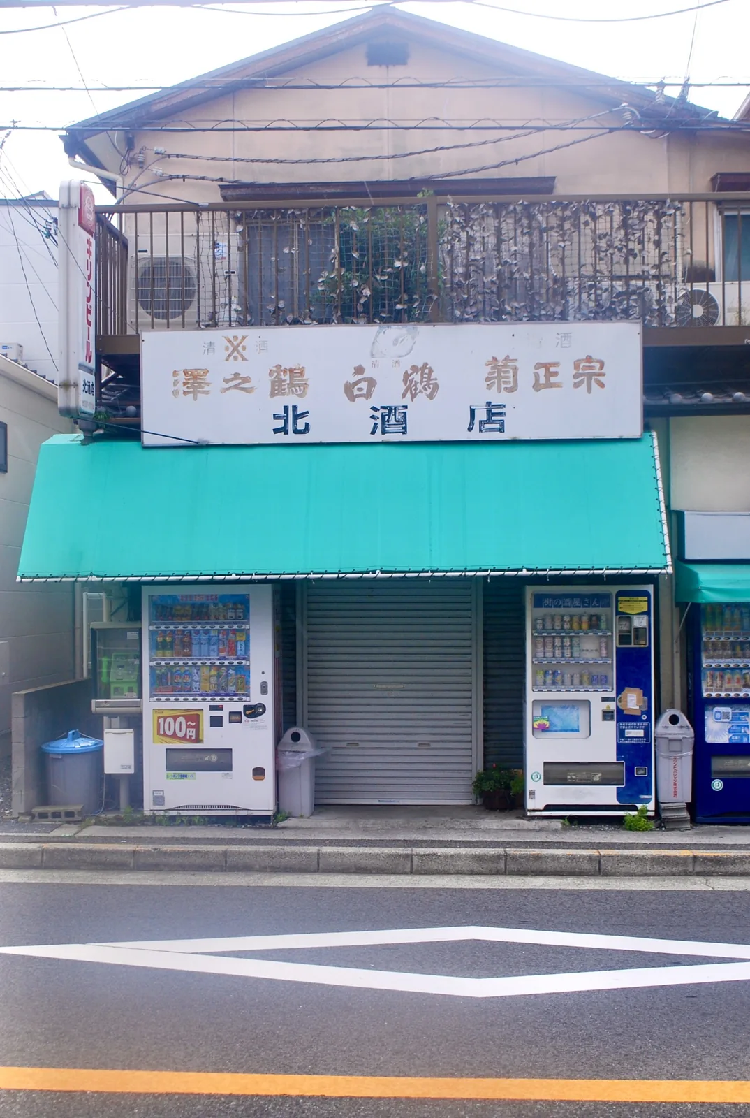 「澤之鶴」「白鶴」「菊正宗」の看板と、緑の日よけ、自動販売機の並ぶ酒店