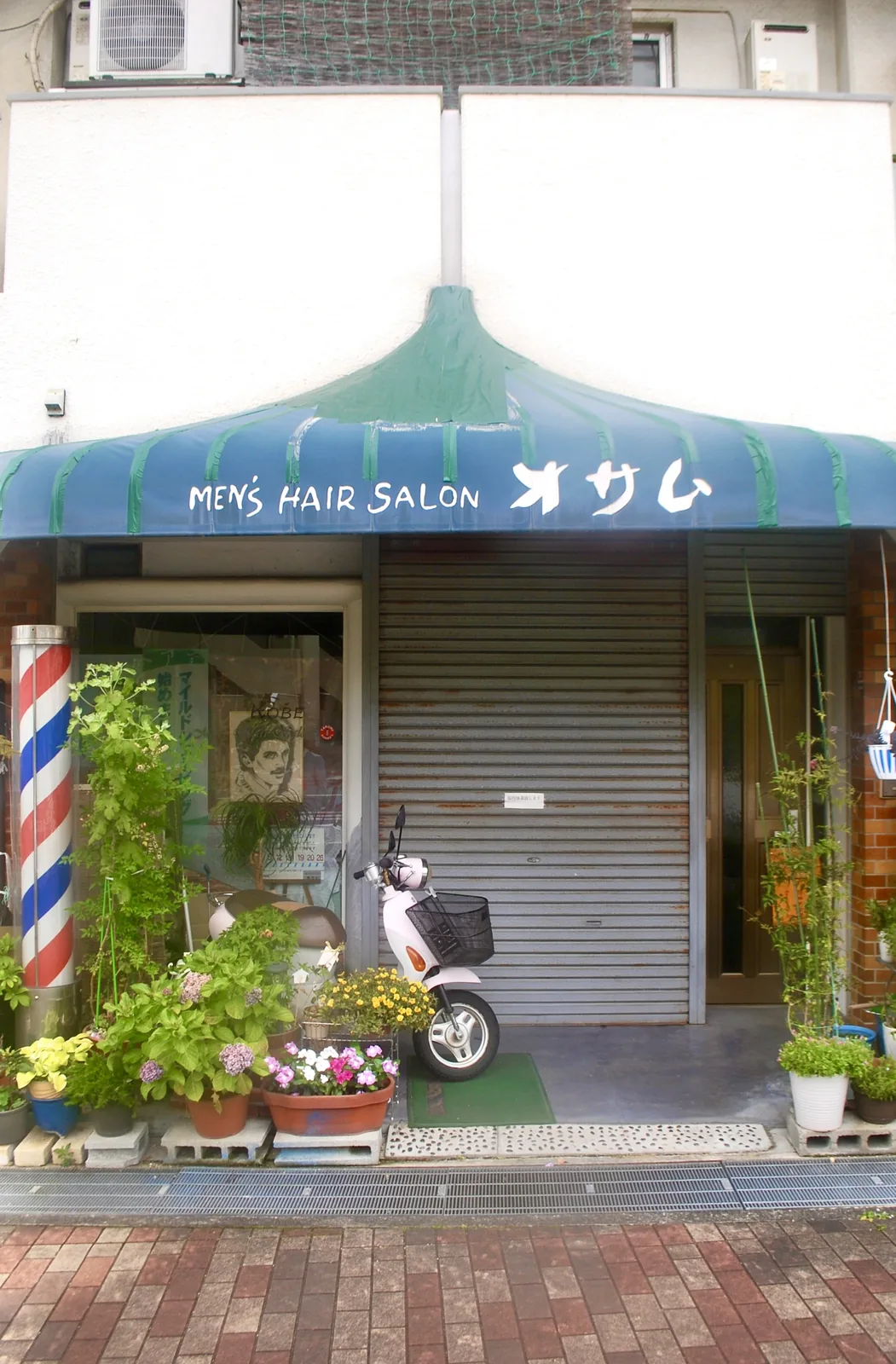 青い日よけとサインポール、花の鉢植えが並ぶ理容店の店先