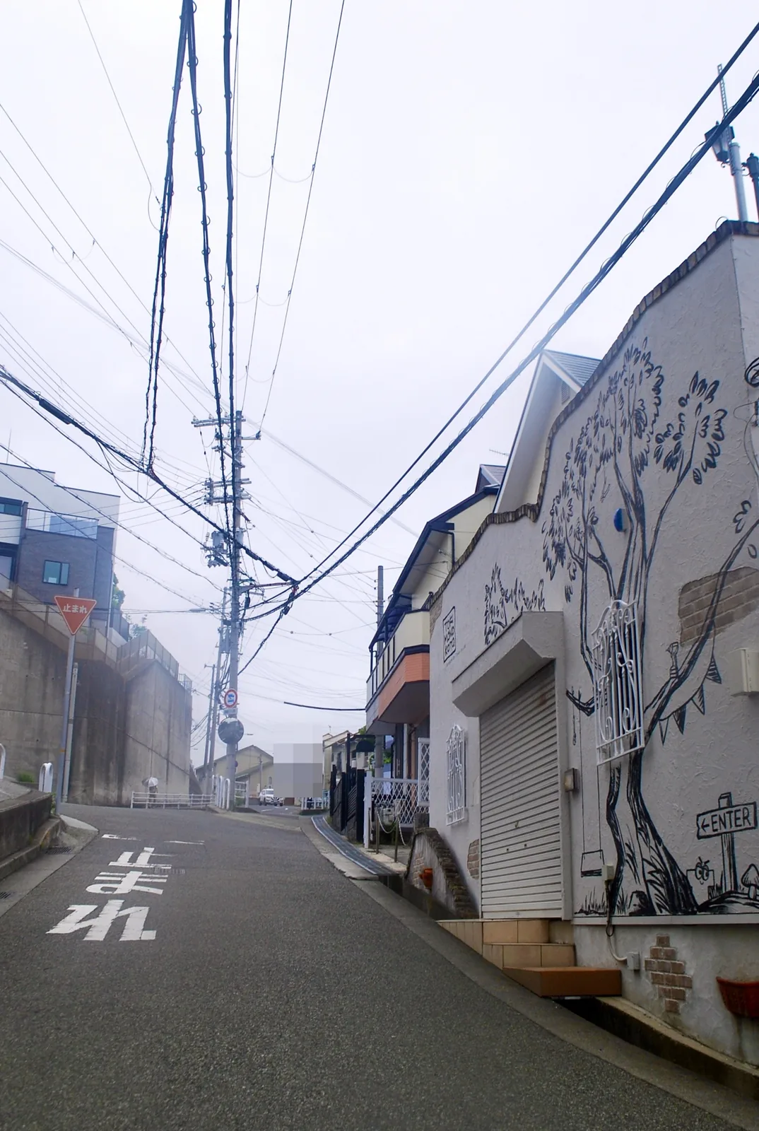大きな木の絵が描かれた家の脇を、上へ続く坂道