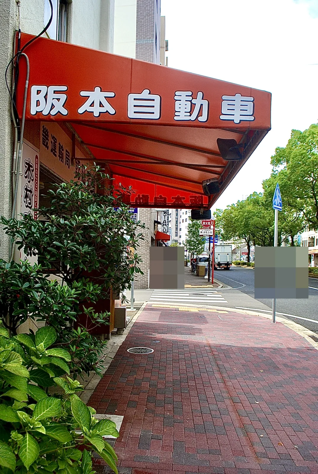 「阪本自動車」の赤い日よけと、植木の並ぶ角の歩道