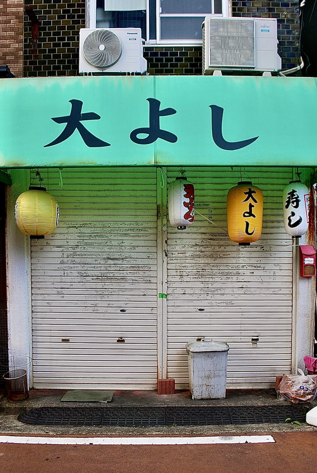 緑の看板の下に、黄色や白の提灯が下がるシャッターの店先
