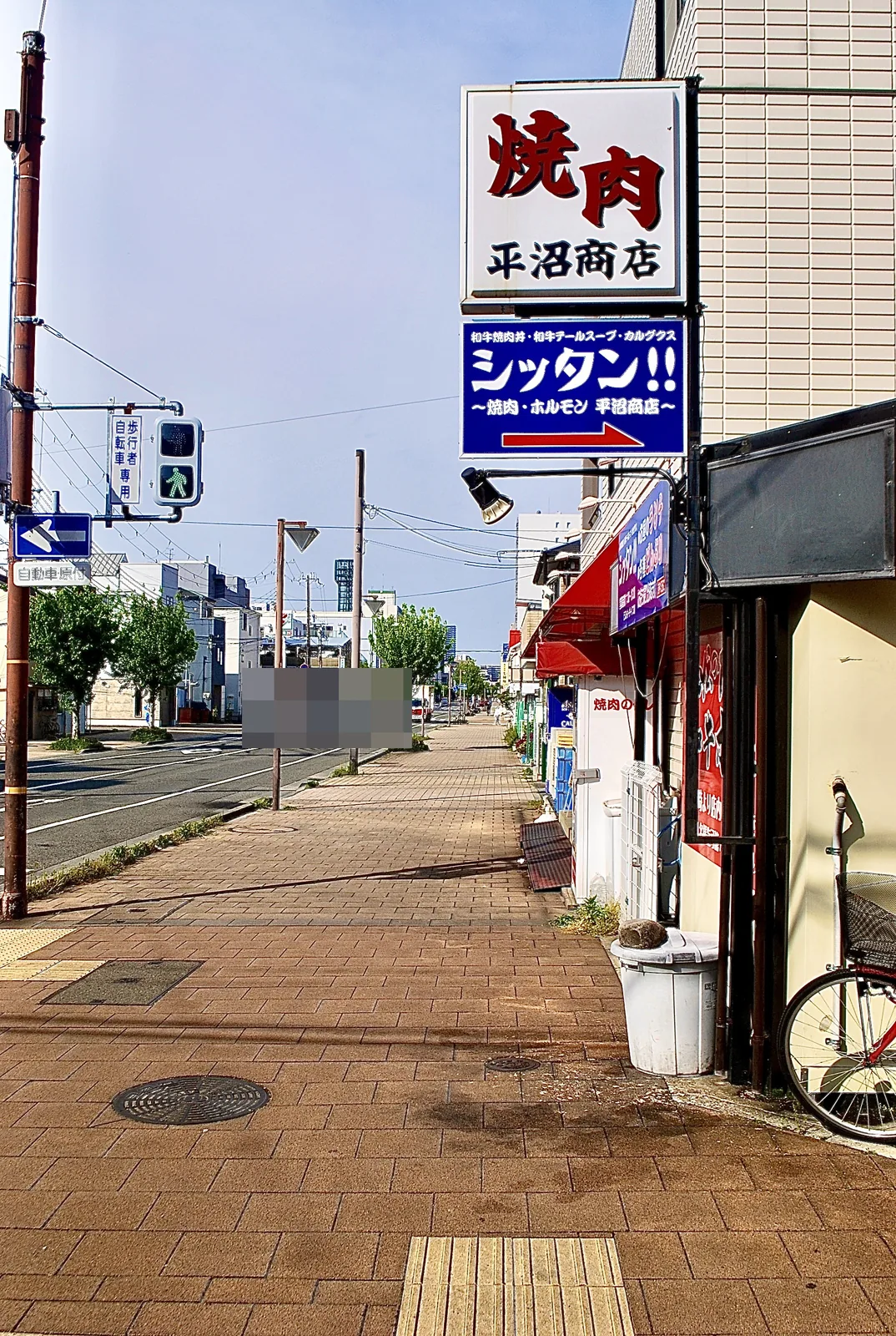 「焼肉 平沼商店」の看板と、並木の続く大通りの歩道