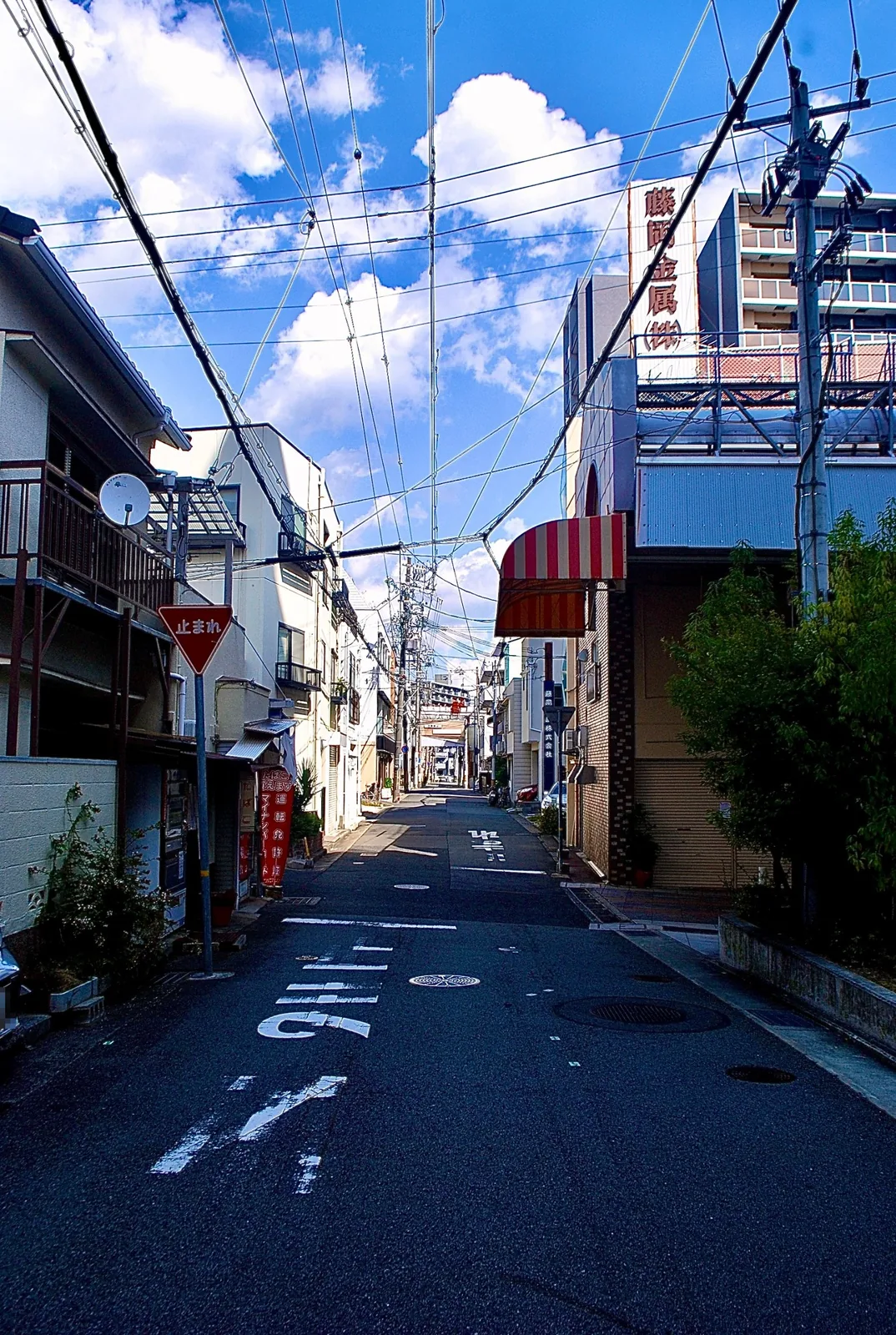 雲の浮かぶ青空に電線が幾重にも張られた、人通りの少ない通り