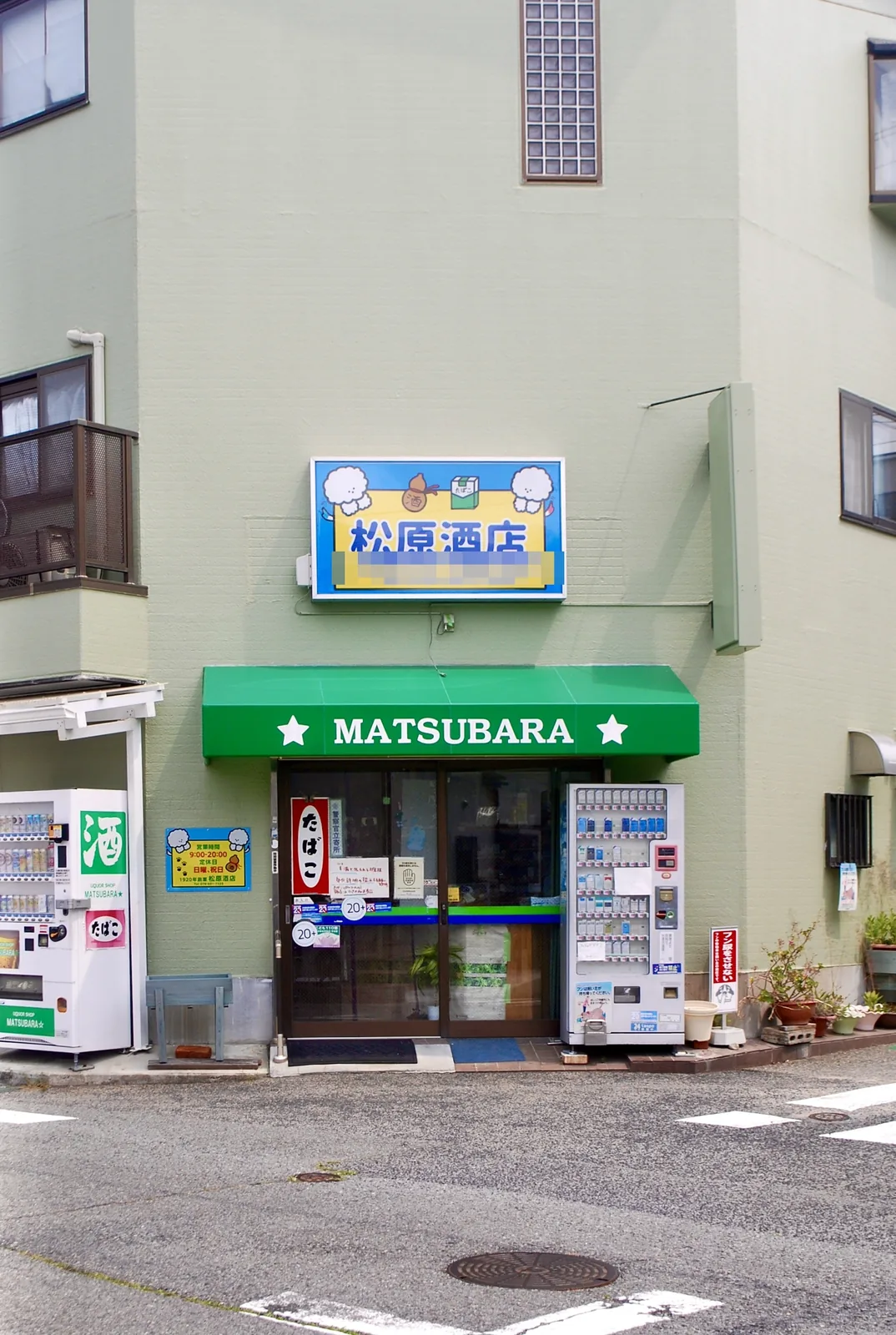 緑の日よけと自動販売機が並ぶ、角の酒店