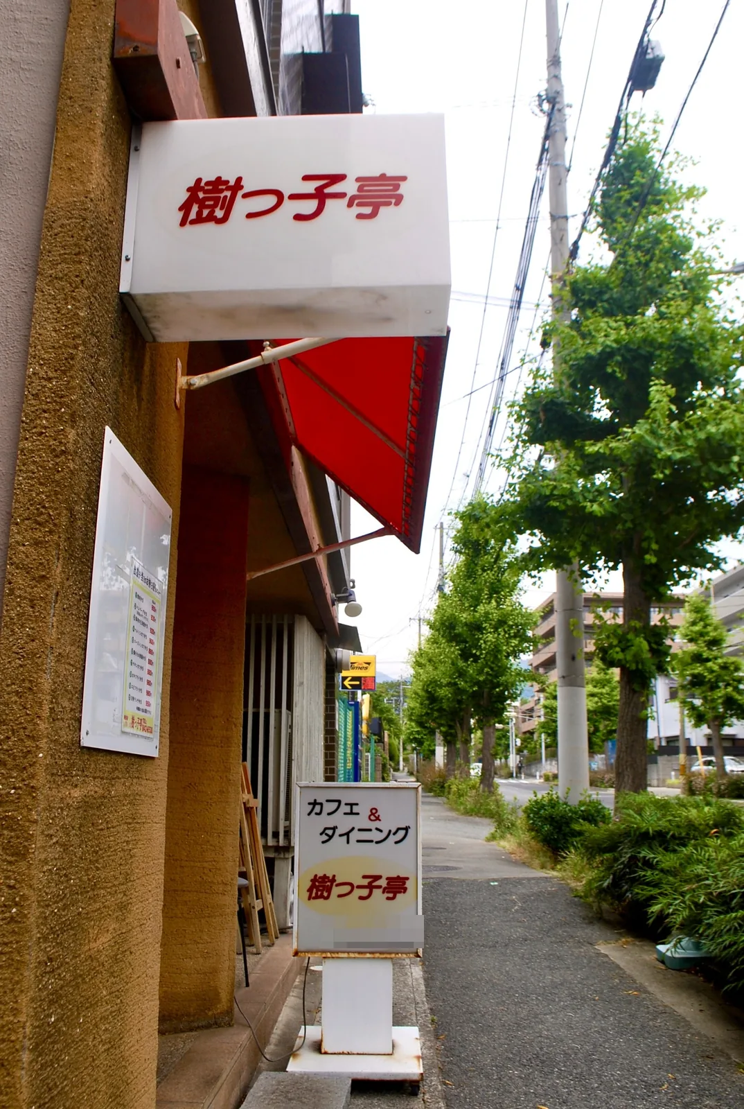 赤い日よけのカフェの横から、並木の続く歩道を見る風景