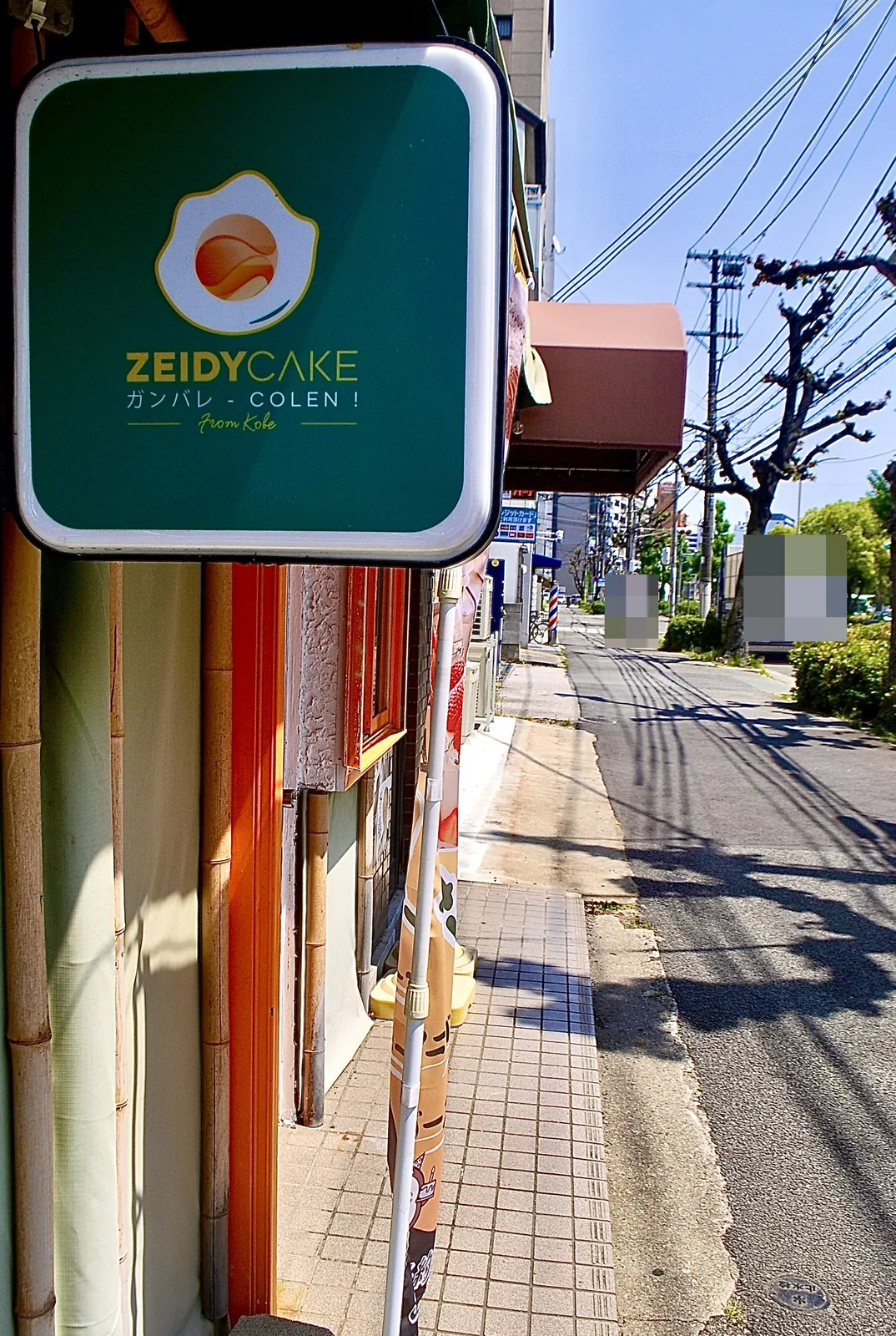 目玉焼きのマークのケーキ店の看板と、並木沿いの歩道