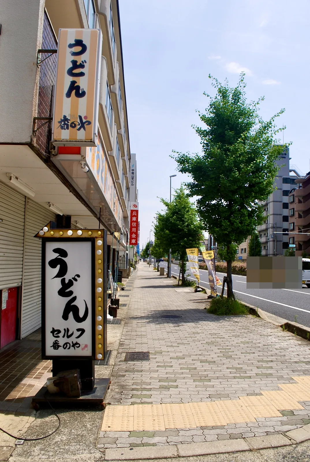 電球に囲まれた「うどん」の看板と、並木の続く大通りの歩道