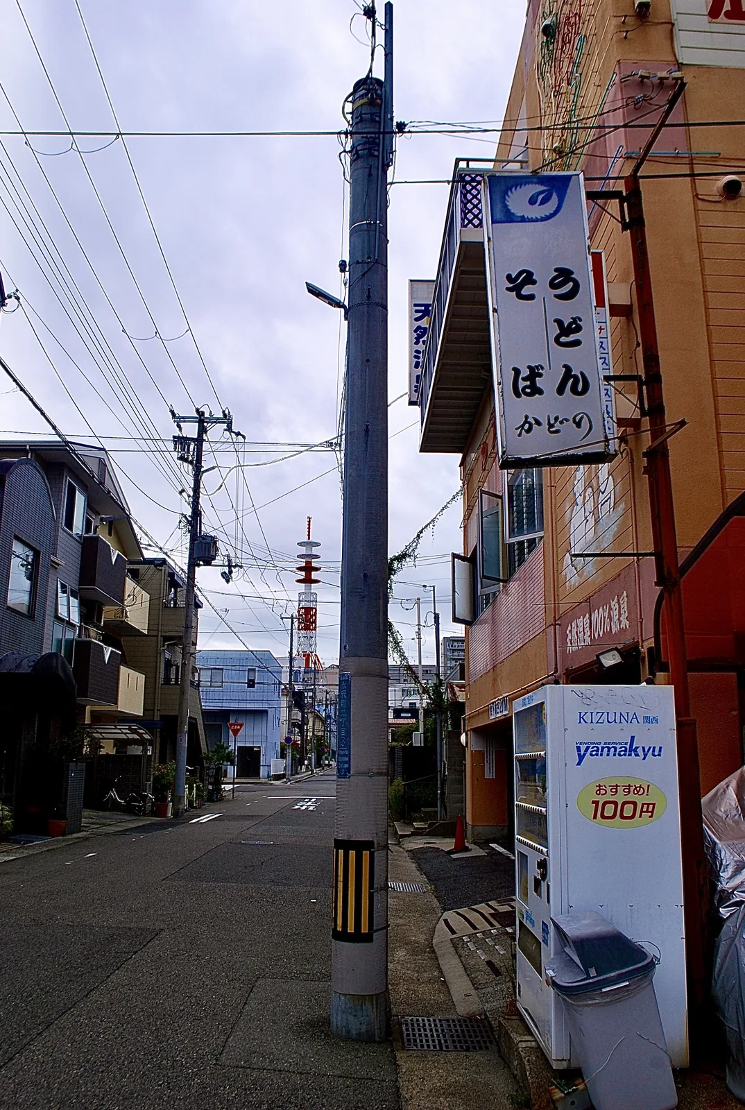 「うどん そば」の看板と自動販売機が並ぶ、細い道