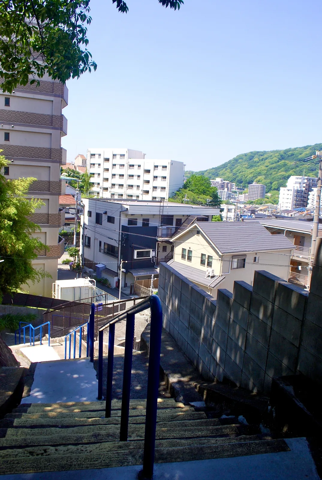 青い手すりの階段の先に、町並みと緑の山が見える風景