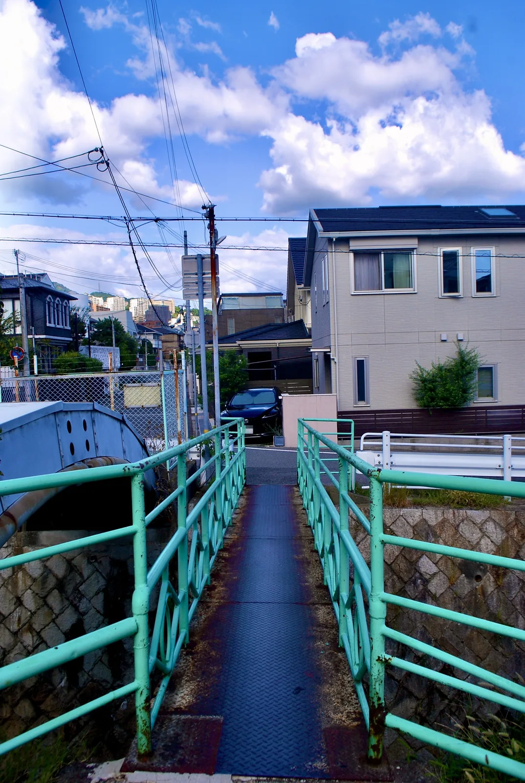 水色の欄干の小さな橋の先に、住宅と青空の広がる風景