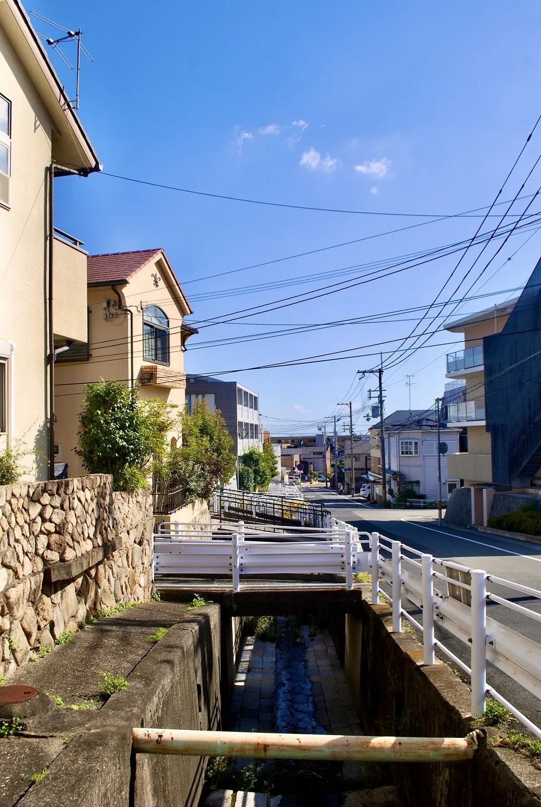 小さな水路に架かる白い柵の橋と、電線の張られた青空の通り