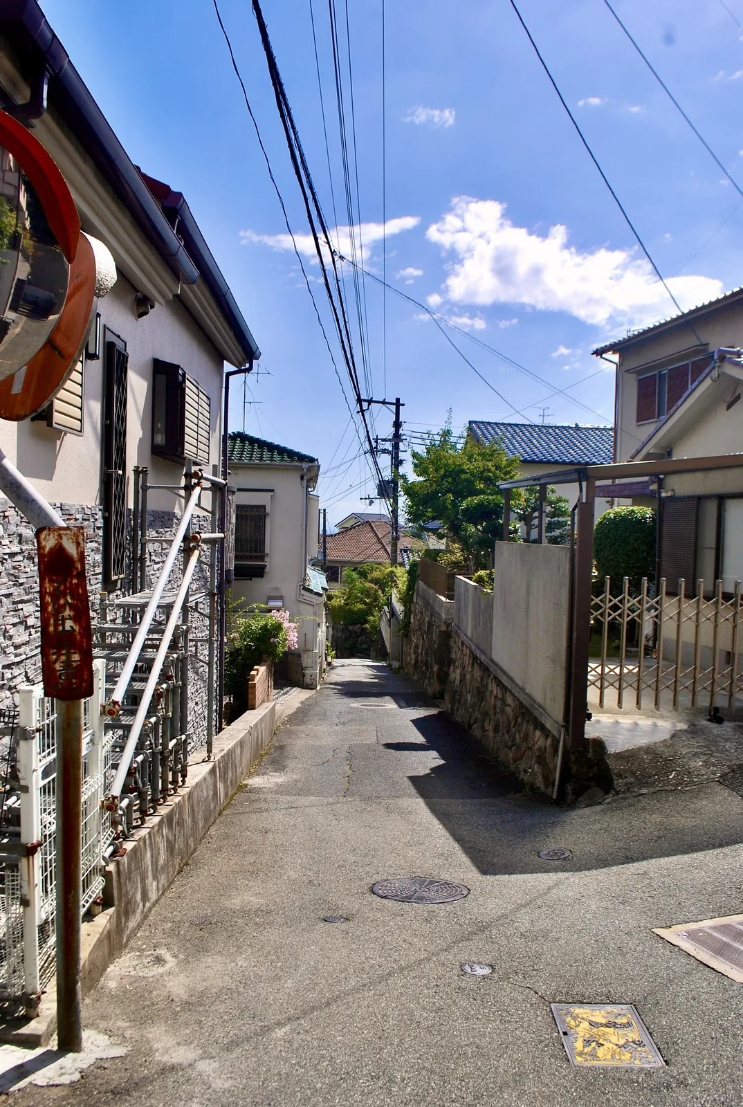 カーブミラーと手すりのある家の脇を、奥へ続く住宅街の路地