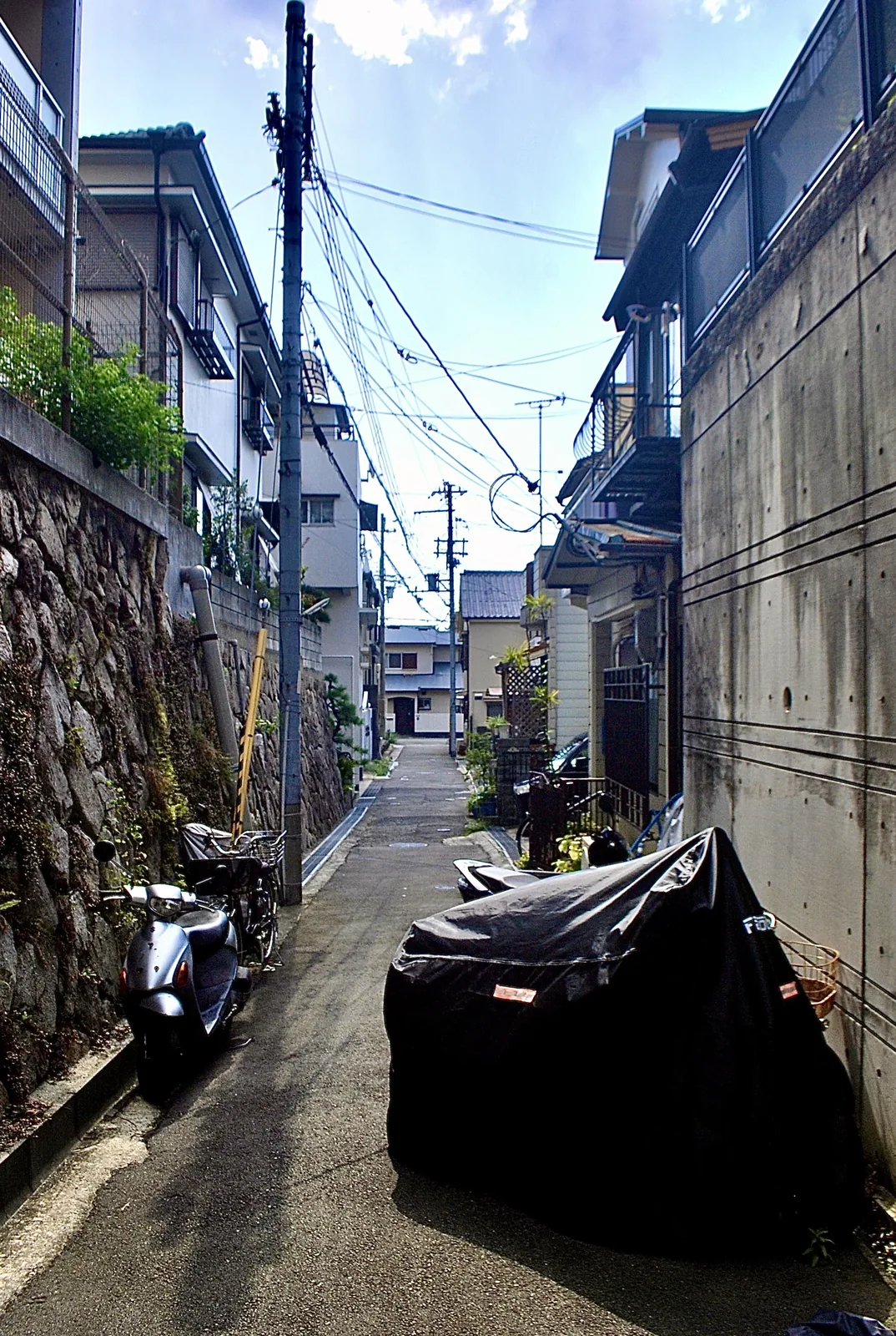 石垣の脇にスクーターが並び、電線の下に続く細い路地