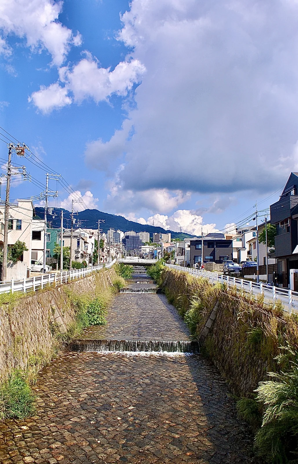 段差のある石積みの川の先に、山並みと町が見える風景
