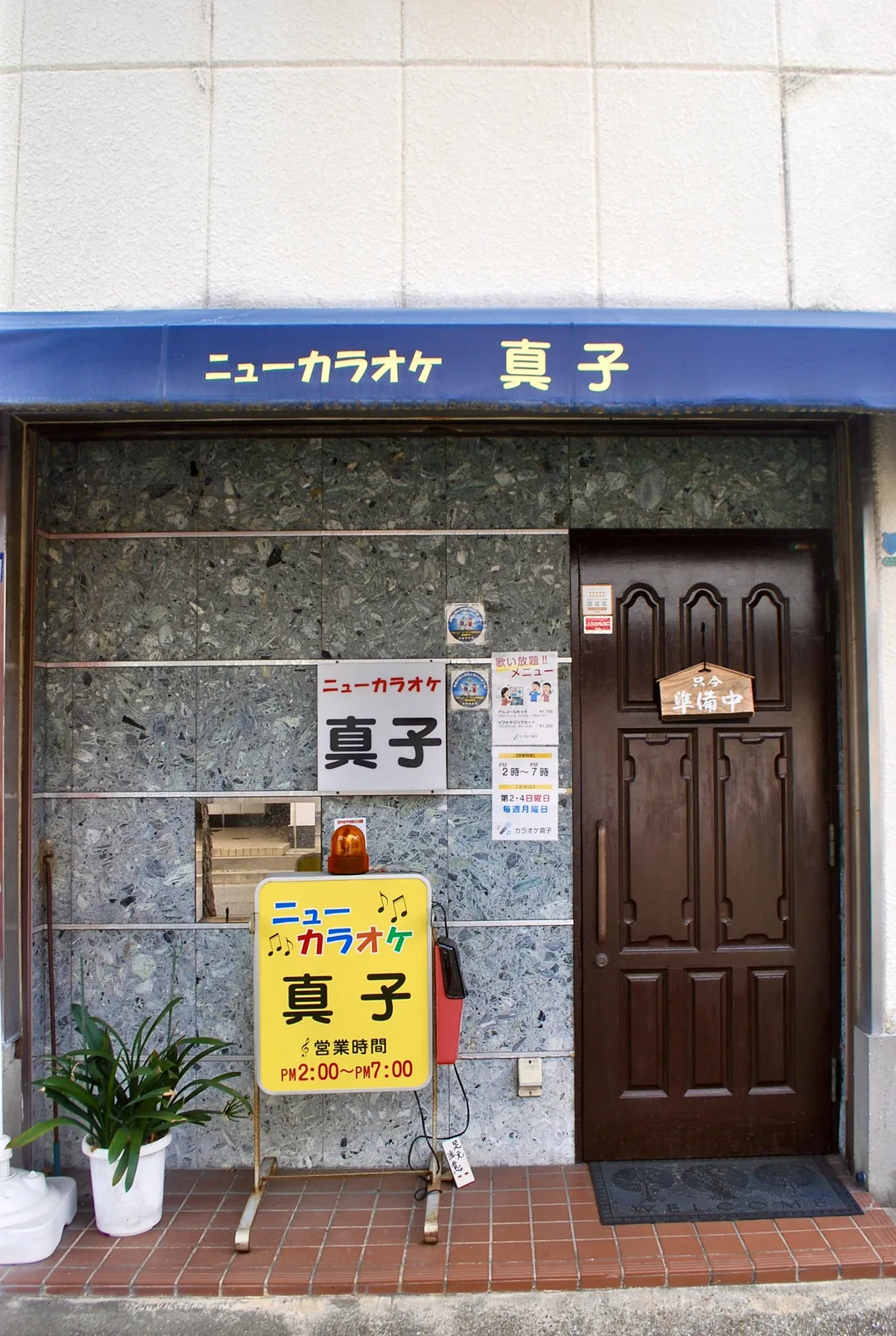 石張りの壁に黄色い看板と木の扉が並ぶ、カラオケのお店
