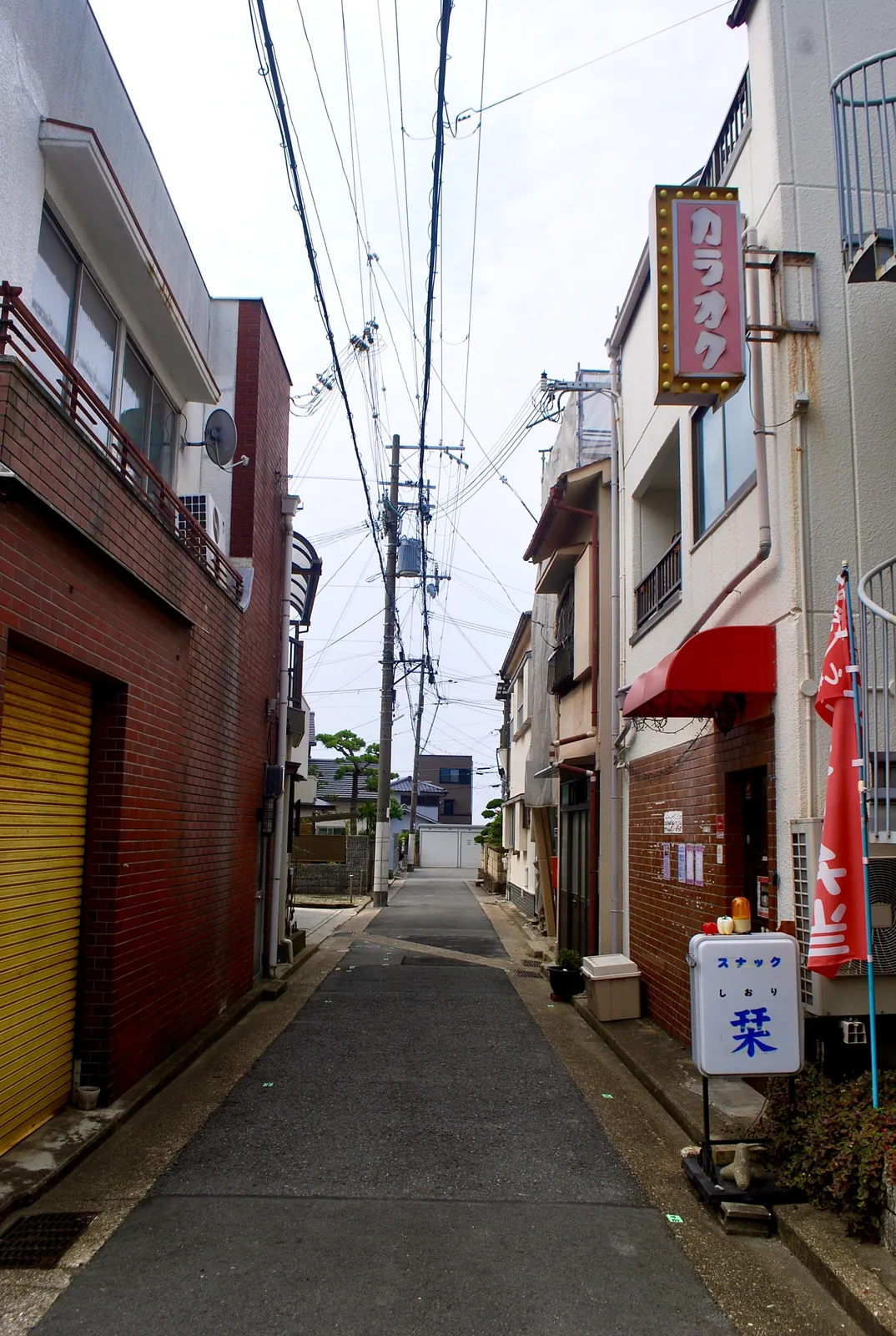 レンガの壁と赤いひさしの先に、スナックの看板が立つ路地